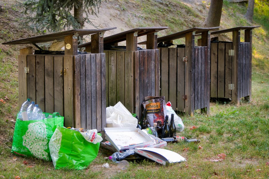 A large pile of black garbage bags filled with waste, stacked against a building with a tiled exterior wall in an outdoor urban area. Some bags are torn open, revealing plastic bottles and miscellaneo